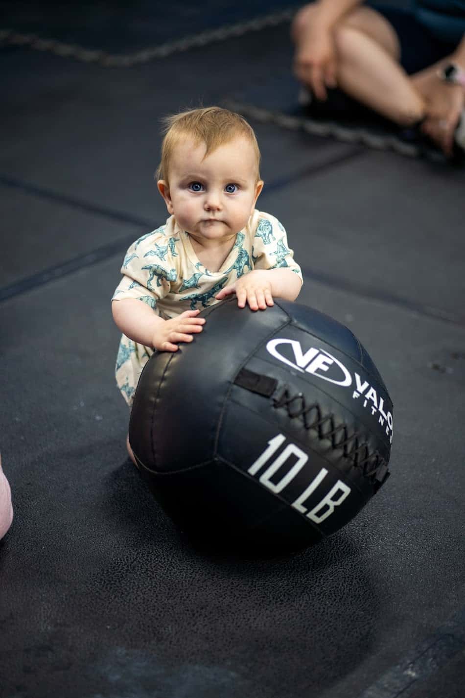 A baby leaning on a wall ball on the gym floor during Diapers and Dumbbells